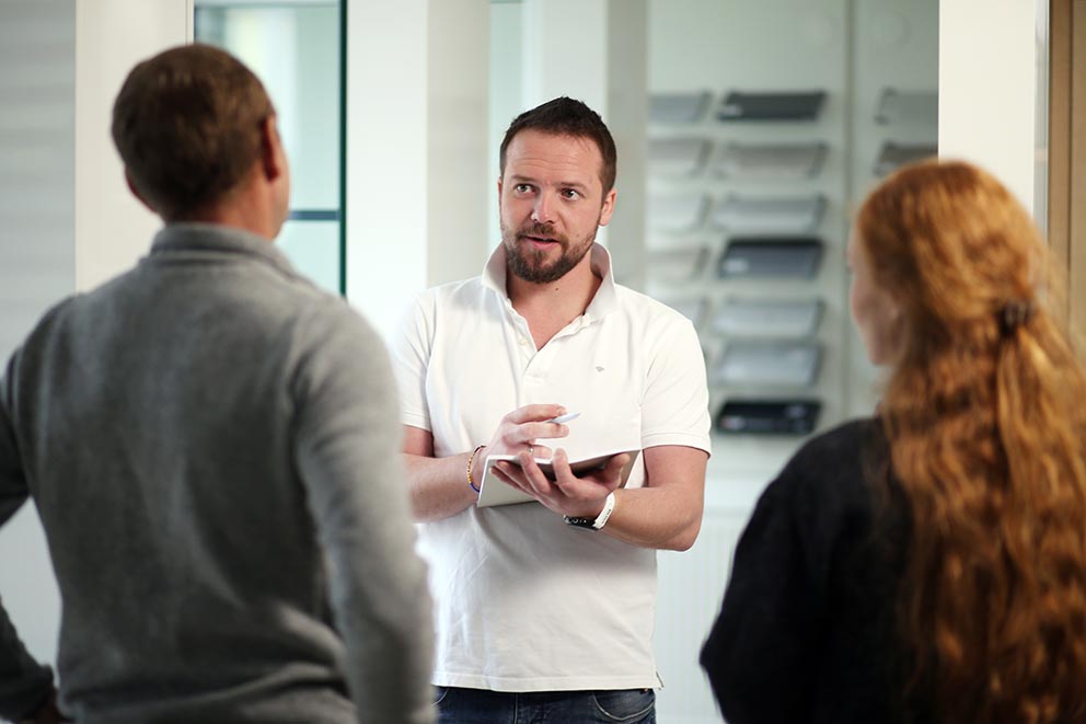Mann in weißem Poloshirt hält Tablet und spricht mit zwei Personen